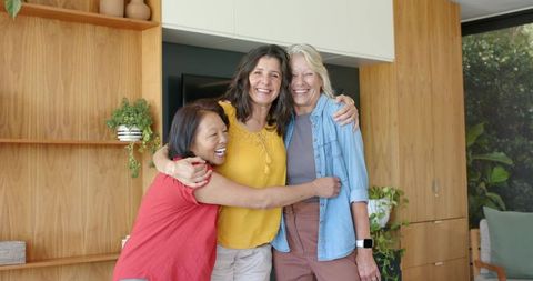 Three women hugging and smiling in cozy modern living room with plants and warm decor