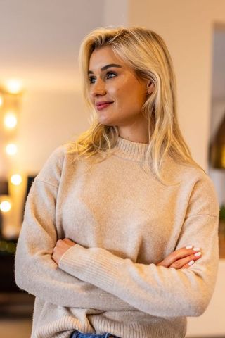 Confident blonde woman standing arms crossed wearing beige sweater in cozy warm interior