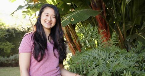 Smiling asian woman touching ferns in sunlit tropical garden with golden star accents