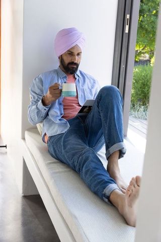 Man in turban relaxing by window with tablet and coffee mug