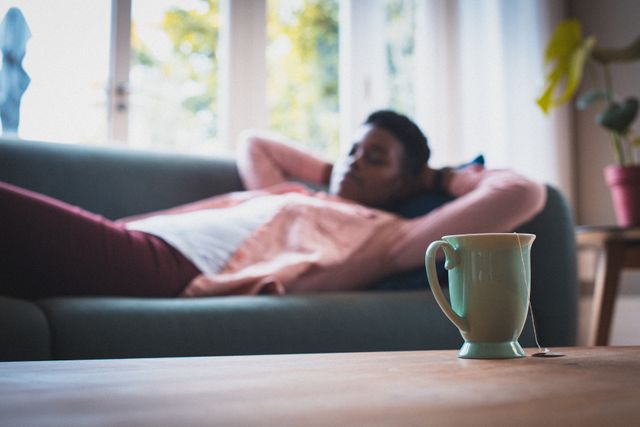 African American woman lying on sofa, relaxing with a cup of tea in the foreground. Ideal for use in articles or advertisements related to home comfort, relaxation, self-care, quarantine activities, and cozy indoor settings.