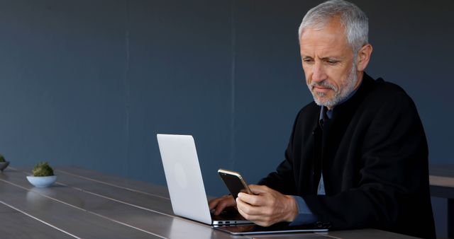 Senior professional with gray hair using smartphone while working on laptop at modern office table. Suitable for business, technology, remote work, corporate, professional life, and office space themes.