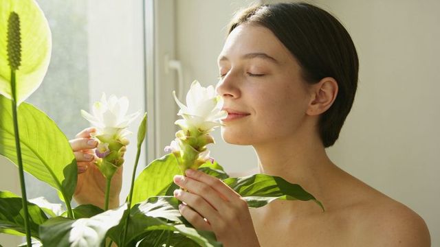 Smelling white tropical blooms at sunlit window, woman cradling curcuma for relaxation