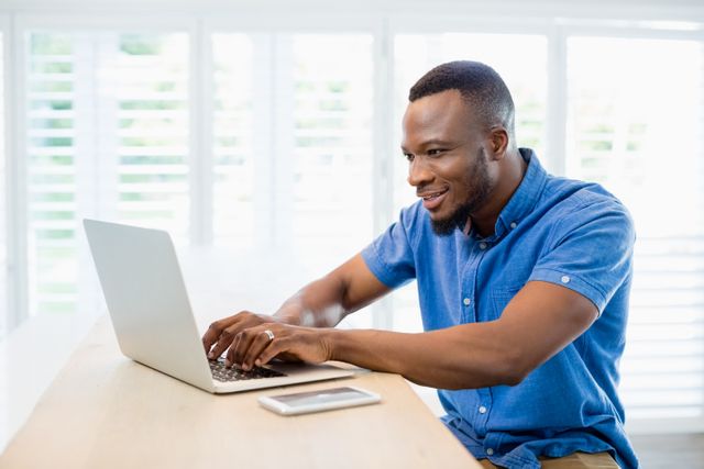 Man sitting at desk using laptop in bright living room. Ideal for illustrating remote work, freelancing, home office setups, or modern digital lifestyles.