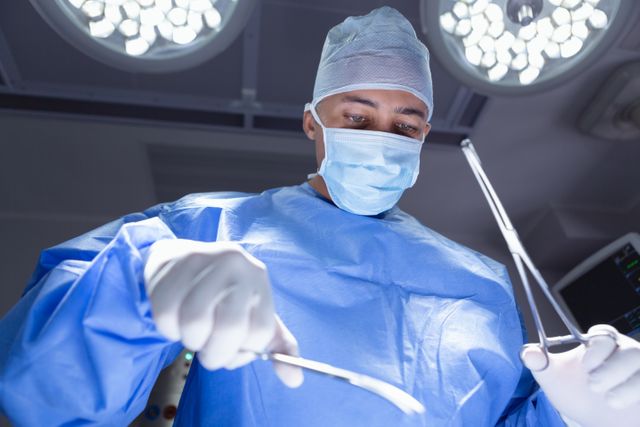 Low angle view of a male surgeon performing surgery in a hospital operating room. The surgeon is wearing blue scrubs, a mask, and gloves, focusing intently on the procedure. This image can be used for medical articles, healthcare websites, educational materials, and promotional content for hospitals and medical institutions.