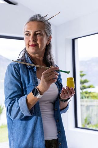 Senior artist painting in sunlit studio wearing denim shirt and smartwatch