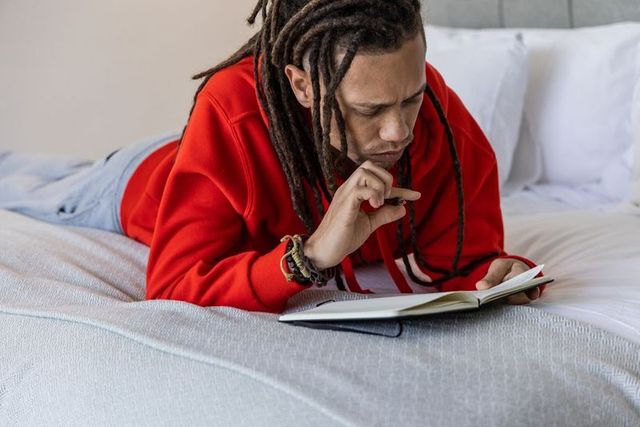 African american man lying on bed reading journal wearing red hoodie and dreadlocks