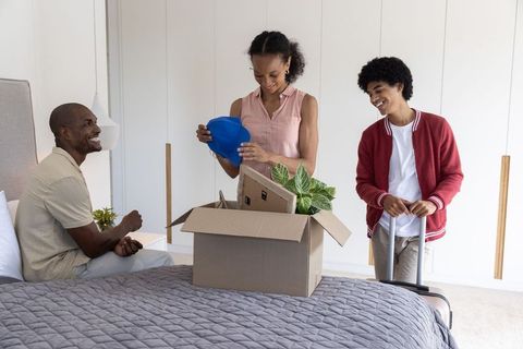 Diverse family smiling, unpacking moving box with blue hard hat in bright minimal bedroom