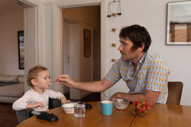 Young father feeding his baby while sitting at the dining table. Strawberries and other breakfast items are on the table. Ideal for use in parenting blogs, family lifestyle articles, advertisements for baby products, or content promoting healthy eating habits for children.