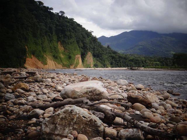 Rock-strewn riverbank stretches into distance with forested hills and mountains in background. Dramatic clouds add to natural beauty and untamed appeal. Ideal for promoting outdoor activities, nature exploration, travel destinations, or nature photography portfolios.