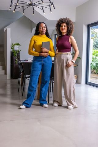 Diverse female colleagues standing in contemporary home office holding tablet