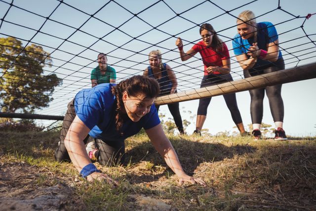 Woman crawling under a net during an obstacle course while her teammates cheer her on. Ideal for use in articles or advertisements about teamwork, fitness challenges, boot camp training, group motivation, and outdoor exercise activities.