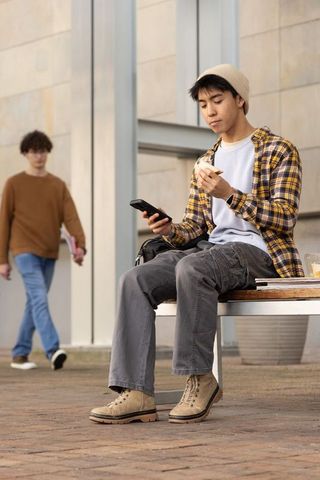 Asian teen using phone while eating sandwich on campus bench, casual student lifestyle