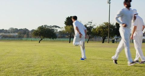 Sunlit cricket teammates warming up with leg-lift drills on grassy field in white kit before match
