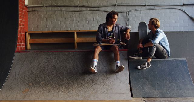 Two friends are having a relaxed conversation while sitting on skateboard ramps in an indoor skatepark. One wearing casual clothes and the other sporting sneakers. This image can be used for themes related to friendship, leisure activities, youth lifestyle, or urban culture. It may be useful for advertisements targeting young adults or while illustrating topics in magazines and blogs focused on skateboarding and casual, active lifestyles.