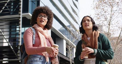 Diverse women holding coffee cups walking outside modern urban campus with backpacks