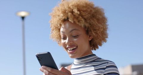 Smiling woman tapping smartphone while reading message in sunny urban plaza