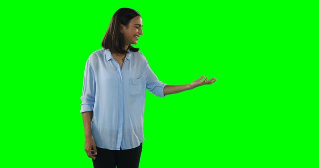 A young Asian woman gestures with her hand as if presenting something, with copy space on the green screen background. Her friendly demeanor suggests she could be a host or presenter in a casual setting.
