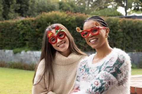 Two friends wearing festive sweaters and novelty red glasses smiling in backyard