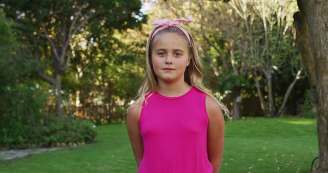 A young girl wearing a pink top and pink headband is standing in a garden, surrounded by greenery and trees. It is a sunny day. She has a confident and calm expression. This image is ideal for use in advertisements, educational materials, family-oriented campaigns, outdoor lifestyle content, and children's fashion catalogs.
