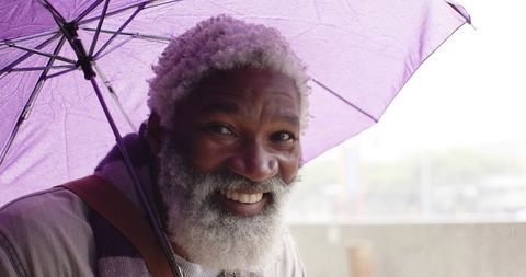 Smiling senior african american man holding purple umbrella in rainy urban street