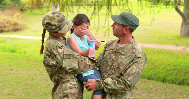 Military parents wearing camouflage uniforms holding and comforting their young daughter in a park. She is holding a small American flag. This visual is powerful for illustrating themes of parental support, military family life, emotional strength, and patriotic values