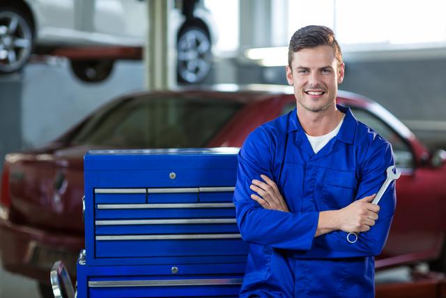 Mechanic in blue uniform standing confidently with arms crossed in an auto repair garage, holding a wrench. Ideal for use in advertisements for automotive services, repair shops, and mechanic training programs. Can also be used in articles or blogs about car maintenance, professional skills, and the automotive industry.