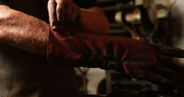 Image shows close-up of worker putting on a protective red glove in a workshop environment. The focus is on safety and preparation. Ideal for illustrating concepts of workplace safety, manual labor, craftsmanship, and industrial job environments.