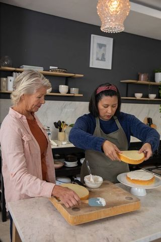 Mature women assembling layered cake in modern kitchen, baking and decorating together