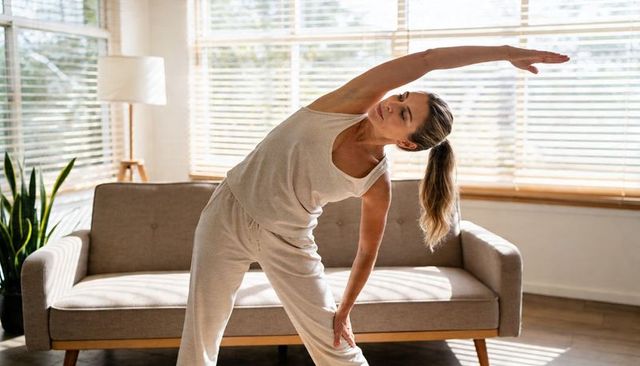 Stretching woman wearing beige loungewear, reaching overhead in sunlit minimalist living room