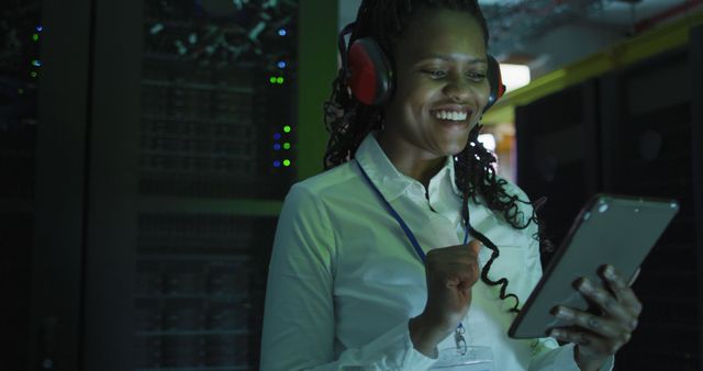 Female IT engineer standing in data center server room, smiling while reading a tablet, wearing headphones and a white shirt with a work badge. This image can be used for illustrating technology jobs, IT professions, data center environments, and professional work settings.