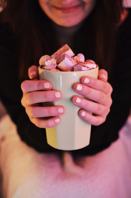 Woman holding a mug filled with hot chocolate topped with marshmallows. Ideal for use in advertisements and promotions related to hot drinks, cozy winter settings, comfort food, or lifestyle content focused on relaxation and indulgence.