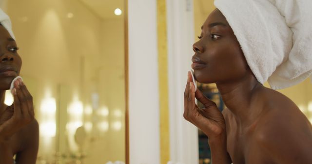African american woman with towel using pad on her face and watching in mirror in bathroom. health and beauty concept.