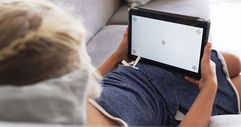 Young woman reclining on sofa holding tablet with blank calibration screen for mockup