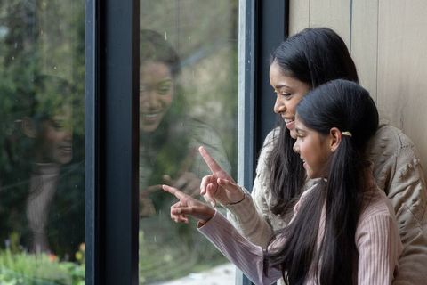 Indian mother and daughter pointing at raindrops through window, cozy indoor bonding