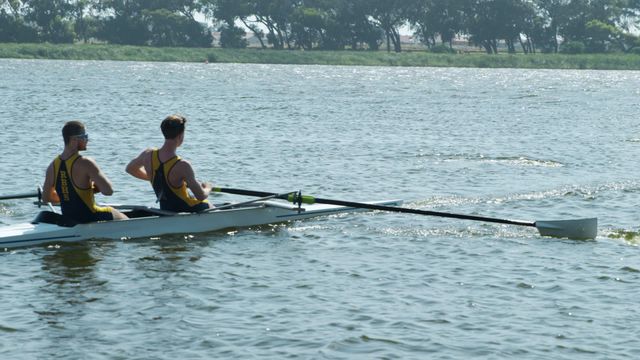 Two teams of Caucasian men are rowing on a peaceful lake, showcasing teamwork and strength on a sunny day. This scene is perfect for promoting team-building activities, sporting events, or fitness campaigns focusing on outdoor exercises and water sports.