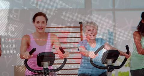 Senior and young adult women pedaling stationary bikes in bright gym class smiling