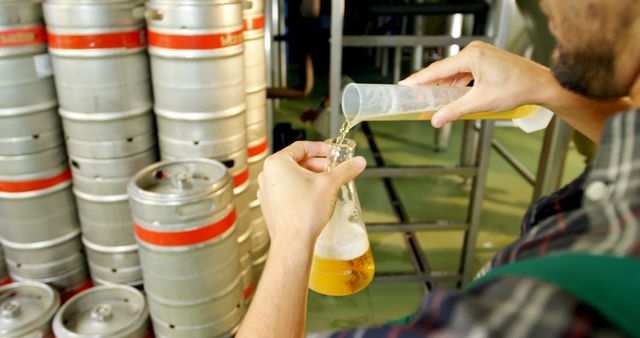 This photo shows a brewer carefully pouring beer into a beaker for quality testing in a brewery environment. Beer kegs line up in the background. The image is useful for illustrating brewing techniques, quality control procedures, brewing industry, and craft beer production in marketing materials, blogs, and websites about brewing.