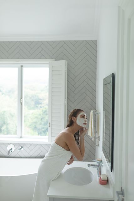Woman wrapped in towel with facial mask looking in bathroom mirror. Ideal for content related to skincare routines, beauty tips, self-care practices, wellness, and home spa experiences. Can be used in articles, blogs, advertisements, and social media posts promoting beauty and relaxation.