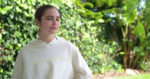Young woman standing in sunlit backyard wearing off-white hoodie and hoop earrings