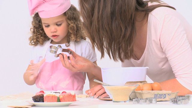 Mother and young daughter baking together, girl wearing pink apron and chef hat. Daughter focused on cookie cutter while mother guides. Perfect for depicting family bonding, cooking instruction, and kitchen activities.