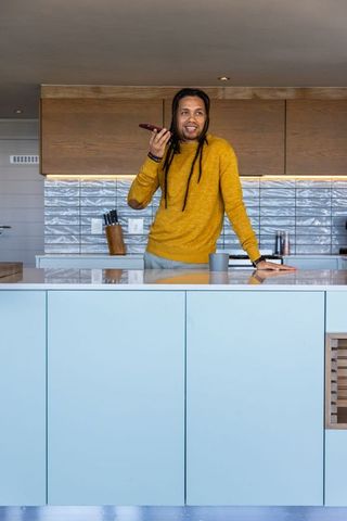 African american man speaking on smartphone at modern blue kitchen island in mustard sweater