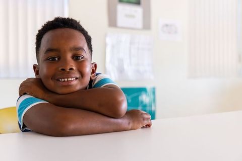 African american boy smiling and leaning on desk in bright classroom for education