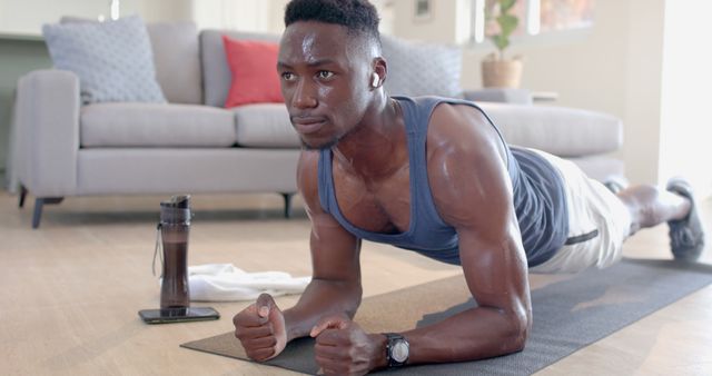 Young man holding plank position on yoga mat in living room. Home workout scene showing determination and fitness. Use for content related to fitness routines, home exercises, and physical health.