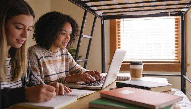 College roommates studying at dorm desk beneath loft bed with laptop and notebooks