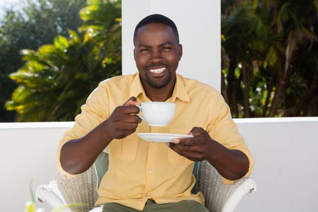 Young man sitting at an outdoor cafe, holding a coffee cup and smiling. Ideal for promoting coffee shops, lifestyle blogs, relaxation and leisure activities, or advertisements focusing on morning routines and enjoyment of nature.