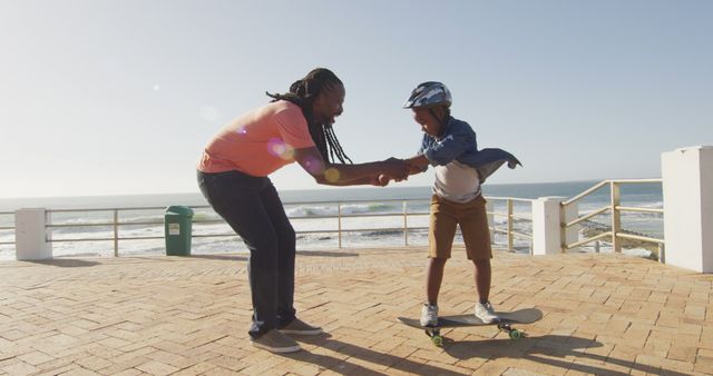 Father helping young son learn to skateboard on a sunny day by an ocean boardwalk. Child is wearing a helmet for safety while gaining confidence and balancing skills under the watchful eye of parent. Perfect for depicting family activities, outdoor fun, and parent-child bonding moments.
