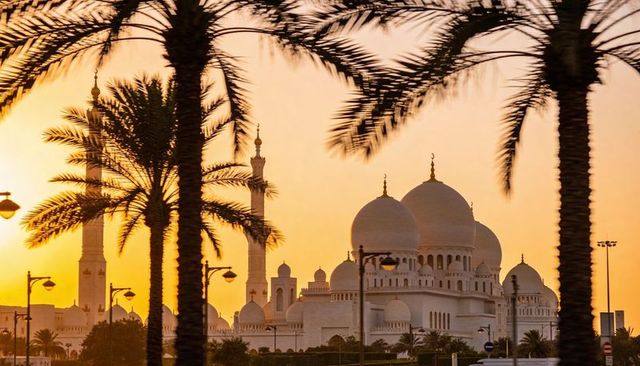Framing grand white mosque and minarets with palm silhouettes, roadside lamps at golden sunset