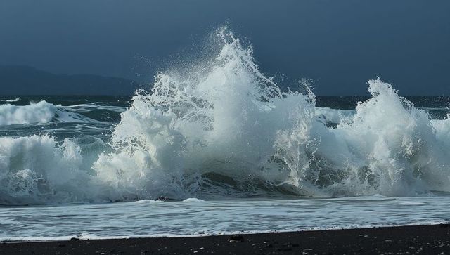 Crashing towering wave exploding into white spray on dark volcanic sand beach