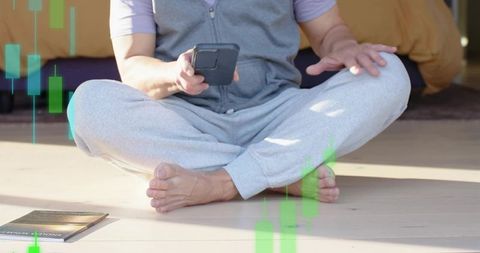 Asian man sitting on bedroom floor using smartphone in gray loungewear, finance overlay